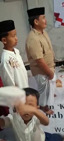 A man in a tan shirt and white sarong stands on a white mat adorned with Islamic symbols and text. He is looking to his right. To his left, a young child in white clothing is partially visible. Behind him, several girls in white hijabs and red skirts stand near a banner. To the right of the man, a group of people, including men and children wearing traditional Islamic hats, are gathered. A man in a teal shirt is holding a rope attached to a bamboo structure. A woman in a red dress and beige hijab is holding a microphone. The environment appears to be indoors, possibly a community center or a hall, with a simple wall and a doorway leading to an outdoor area where motorcycles are parked. The overall atmosphere suggests a gathering or event.