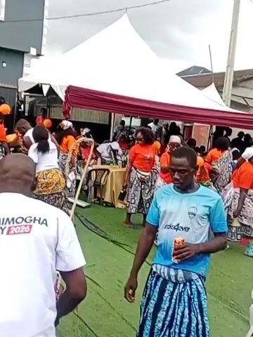 The video appears to capture a lively outdoor event, possibly a celebration or gathering. A large white tent with red drapes is set up, covering an area with green carpeting. In the foreground, a man wearing a blue t-shirt and patterned shorts is holding a beverage and looking towards the camera. Behind him, several women are dressed in matching orange t-shirts and patterned skirts, some of them dancing. Other individuals are visible in the background, some seated at tables and others standing. The overall atmosphere seems festive and energetic.