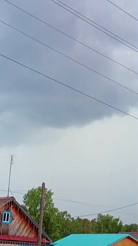 The video shows a view of houses and trees against a cloudy sky. There are power lines crisscrossing the frame. A wooden house with red and white trim and a blue-trimmed window is visible on the left. To its right, another house with a teal-colored roof is partially seen. A wooden pole stands in front of the houses. The sky is overcast with dark clouds suggesting an impending storm.