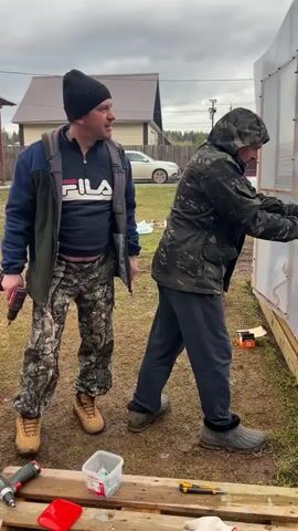 Two men are working on the side of a greenhouse in a muddy backyard. The man on the left, with a black beanie, black jacket with a Fila logo, and camouflage pants, is holding a red cordless drill and watching the other man. The man on the right, wearing a camo jacket with the hood up and dark sweatpants, is kneeling and using a utility knife to trim a clear plastic panel. There's a box of screws and another drill on a wooden pallet. A white SUV is parked in the distance near a house.