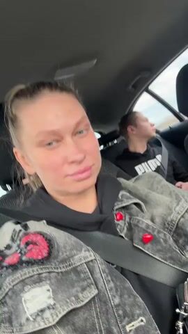 The video captures three people inside a car driving during the daytime in Kitsjanovo, Russia. The person filming from the front passenger seat is a woman with light blue eyes and light brown hair tied back, wearing a black hoodie under a dark distressed denim jacket with red heart-shaped buttons and embroidered details. In the back seat, a boy wearing a black Puma hoodie is visible, looking out the window. The camera then pans to the driver, a blonde woman wearing sunglasses and a brown bomber jacket, with her hands on the steering wheel and a cup in one hand. A small black bag with the word "GUESS" is briefly visible. The car appears to be on a road or highway, with an open landscape visible through the windows under a bright, possibly overcast, sky.