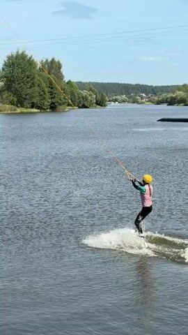 A person in a yellow helmet and a pink and blue life vest is wakeboarding on a lake. They are holding onto a rope that is attached to an overhead cable system. The person is creating a wake behind them as they move across the water. Trees and a blue sky with white clouds are visible in the background.