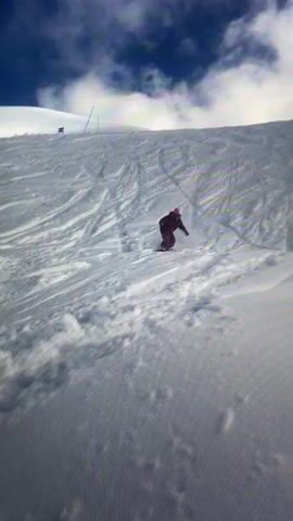 A person is seen snowboarding down a snow-covered mountain slope on a cloudy day. They are wearing a red jacket and a pink hat. Initially, the snowboarder is upright, maneuvering down the slope. Suddenly, they lose balance and fall backward onto the snow. The impact causes their pink hat to fly off their head and land a short distance away.

The scene then widens to show the extensive ski area. A ski lift with multiple chairs is visible overhead, spanning across the slope. The ground is covered in fresh snow, crisscrossed with numerous ski and snowboard tracks. In the background, majestic snow-covered mountains rise, with some peaks partially obscured by clouds. Bare tree branches are visible in the immediate foreground of the wider shot, adding to the winter landscape. The fallen snowboarder and their detached pink hat remain visible on the slope. The weather appears to be daylight with mixed cloud cover. There is no visible text in the video. The location context provided is Villeurbanne, France.