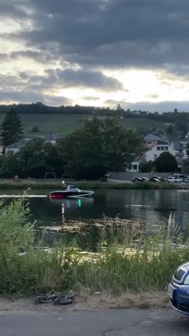 A motorboat with red and black accents and a green light on its side travels across a body of water. The boat is moving from left to right in the frame. Beyond the water, there is a green hillside dotted with houses and trees. The sky is cloudy with patches of bright light, suggesting a sunset or sunrise. In the foreground, tall green grass and a portion of a blue car are visible. The scene appears to be a calm, outdoor setting.