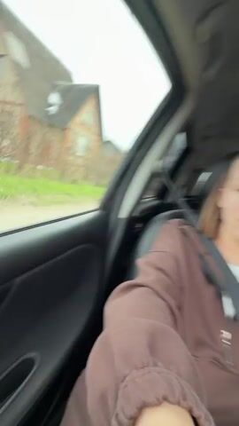 A woman is taking a selfie video from the passenger seat of a car while a man is driving. Both are wearing similar brown zip-up sweatshirts and seatbelts. The woman has light hair and is looking directly at the camera while talking. The man driving has a shaved head and is looking straight ahead. In the backseat, there are various items including what appears to be a white box and a black object. The car is moving, as indicated by the changing scenery visible through the windows. The lighting is bright and natural.