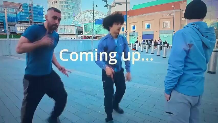 Two young men are standing on a paved area in front of a modern urban background. The man on the left has a beard and is wearing a blue t-shirt and black pants. He is gesturing with his hands and appears to be speaking or rapping. The man on the right has curly dark hair and is wearing a grey zip-up jacket and black pants. He is also gesturing with his hands. Text overlay on the video reads "Coming up...". The background includes buildings with glass facades, scaffolding on one building, streetlights, and bollards. Other people are visible in the background, walking and standing. The overall impression is that of a dynamic urban setting, possibly a street performance or a preview for an event.