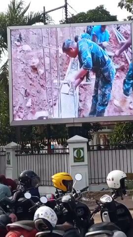 A digital billboard displays a video of several people in military uniforms performing construction work outdoors. The construction scene shows a muddy area with rebar and concrete being poured into forms. In the foreground, a row of parked motorcycles with helmets on them is visible. The billboard is mounted on a pole, and behind it are trees and a building. There are also two ornate pillars with a circular green emblem in front of the billboard.