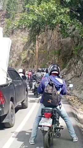 The video shows a traffic jam on a road in San Antonio de Los Altos, Venezuela. The primary subjects are people riding motorcycles and a black pickup truck filled with various household items. The pickup truck is loaded with a large blue barrel, stacked cardboard boxes, wooden planks, and what appears to be a mattress or large piece of furniture. The motorcycles are mostly carrying two people, with riders and passengers wearing helmets.

The scene is outdoors on a paved road, with a steep, tree-covered hillside to the right and a clear sky with scattered clouds suggesting daytime. The weather appears to be sunny and warm. In the background, there are buildings with palm trees, indicating a developed area.

The activity taking place is a traffic jam, with vehicles moving slowly or stopped. The loaded pickup truck and the group of motorcycles suggest a possible move or transport of goods, or people traveling together.

Notable details include the license plate on the pickup truck, which reads "AH949E", and the license plate on one of the motorcycles, which reads "JU949E". The rider in the foreground on the motorcycle wears a blue hoodie and a black backpack. The overall mood seems neutral, typical of a traffic situation. The visible text includes brand names on the boxes in the truck, such as "PREMIUM".