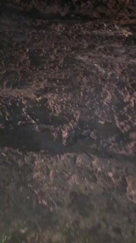 The video shows a muddy, uneven surface, likely a dirt road or trail. Tire tracks are visible in the mud, suggesting that a vehicle has recently passed through the area. There are also some patches of what appears to be water or wet mud, indicating recent rain or damp conditions. In the upper right corner of the frame, some green vegetation is visible, suggesting an outdoor environment. The lighting is dim, with some areas illuminated by what appears to be artificial light, possibly from a flashlight or headlights. The camera is moving, as evidenced by the slight blur and the changing perspective, indicating that someone is walking or driving through the muddy terrain. There is no visible interaction between people or any specific objects of interest beyond the general environment.