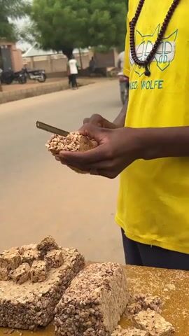 A man in a yellow t-shirt cuts a block of peanut brittle with a knife. He is standing next to a table on which there are three large blocks of the brittle and some smaller pieces. A street and other people are in the background.