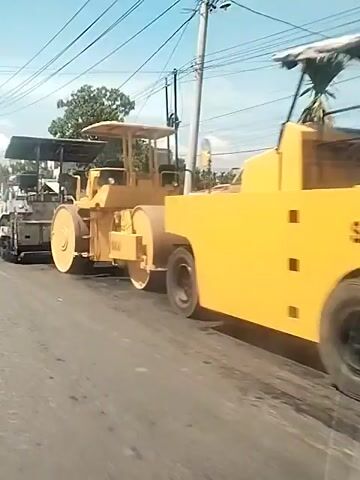 The video depicts road construction activities. Initially, a yellow Sakai-branded road roller is visible, followed by a paving machine. The camera then pans to follow an orange dump truck moving on the road. The environment includes overhead power lines, trees, and buildings along the roadside.