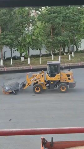 A yellow wheel loader equipped with a street sweeping attachment is cleaning a paved surface. The loader is in motion, and the sweeping mechanism is actively rotating, kicking up dust. The scene is set outdoors, likely on a street or parking lot. In the background, there are trees with green foliage, a white building with a fence, and white posts with red tops, suggesting an urban or suburban environment. The sky is not visible, but the lighting indicates it is daytime. There is no visible text on the loader or its attachment, except for the model number "542" and the brand "ELGI" on the loader's arm. The overall atmosphere appears to be one of routine maintenance.