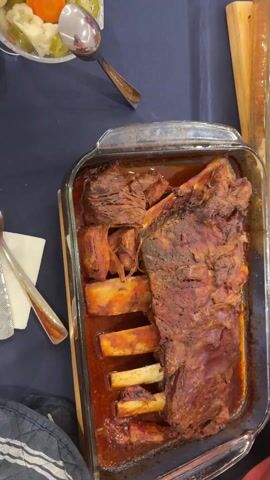A top-down view reveals a glass baking dish filled with cooked beef ribs. The ribs are arranged in a reddish sauce, with some meat shredded off the bone. To the left of the dish is a folded napkin and silverware. In the top left corner is a bowl of pickled vegetables with a spoon. A portion of another dish is visible on the right. The scene appears to be a table setting.