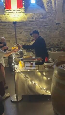 The video begins by showing a food service area decorated with fairy lights, set up against a stone wall. A person in a black shirt and cap is handing a plate of food to another person. In the background, more people are seated at tables in a dimly lit, vaulted stone chamber. Wine barrels are visible, contributing to the rustic ambiance. The camera pans to reveal the interior of the chamber, showing more tables and patrons. The scene suggests a casual dining establishment, possibly a restaurant or event space with a historical or medieval theme.