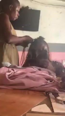 A woman in a tan dress is braiding hair in a room. The hair is long, brown, and flowing. The woman braiding the hair is holding the hair with both hands, and her face is visible in profile. A person is seen reflected in the mirror, holding a cell phone. The surrounding environment includes a table covered with a pink cloth, a mirror, and walls of a light color. A flat screen television can be seen on the wall in the background.