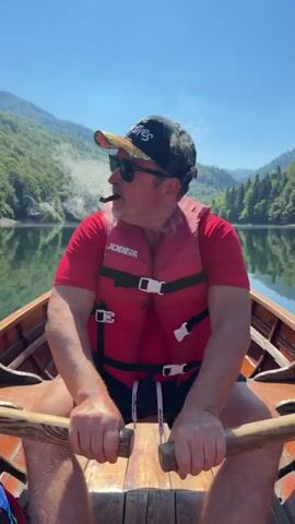 A man is sitting in a wooden rowboat, holding oars and rowing. He is wearing a red t-shirt, black shorts, a red life vest, sunglasses, and a baseball cap. He is smoking a cigar. The boat is on a lake, with mountains and trees in the background. The sky is blue. The video is shot from the perspective of someone sitting behind the rower.