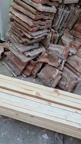 The video shows a collection of old, weathered red roof tiles stacked on the ground next to a pile of new, light-colored wooden planks. The tiles are rectangular, with curved edges and a raised pattern on the surface. Some of the tiles are cracked or broken, and they appear to be covered in dirt and moss. The wooden planks are neatly stacked, and they appear to be freshly cut.

The ground is mostly concrete, with some patches of dirt and grass. In the background, there is a fence, a wall, some greenery, and a vine-covered pergola. The sky is visible above the pergola, and it appears to be a clear day. There is no visible person in the video.