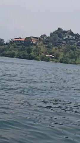 The video shows a body of water with small waves, likely a lake. On the horizon, there's a hilly area with trees and houses. The sky is overcast, creating a muted light over the scene. The camera seems to be on a boat or moving object on the water, indicated by the movement of the waves. There are no people visible in the shot.