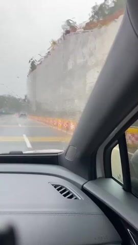 The video, taken from inside a vehicle, shows a view through the windshield and side window on a rainy day. The windshield is covered in raindrops, distorting the view of the road ahead. The road appears to be wet, with yellow and white lane markings visible. Along the side of the road, there are orange and white traffic barriers. In the distance, there are a few red taillights of other vehicles. To the right, a portion of the car's interior is visible, including the dashboard and a speaker. The side window shows a large, textured concrete wall. The weather is overcast and gloomy.
