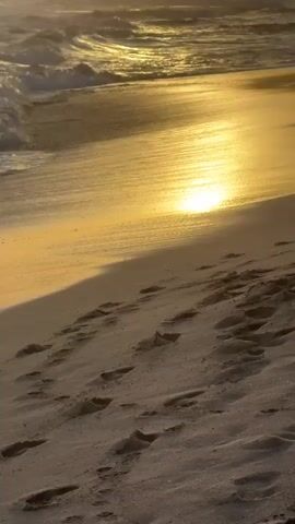 The video captures a serene sunset scene on a sandy beach. Golden light from the setting sun, partially obscured by clouds, casts a warm glow over the ocean and shoreline. Waves gently roll onto the beach, with the sunlight reflecting brightly on the wet sand and water. Footprints are visible in the dry sand on the right side of the frame. In the distance, silhouetted palm trees and a building are visible against the bright sky, enhancing the tropical atmosphere. The scene is tranquil, depicting the natural beauty of the coast at dusk in Thalaramba, Sri Lanka.