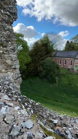 The video presents a scenic outdoor view, captured from atop weathered stone ruins. In the immediate foreground, the rugged surface of the ruins is visible, composed of small stones, gravel, and patches of moss and dirt. Beyond this, a sloping, vibrant green grassy area descends towards a cluster of trees and a building.

The building, located in the middle ground, features a combination of reddish brick and lighter stone work, with white arched windows and a distinctive green door. Its grey roof is equipped with solar panels. The building is enveloped by various trees, some with bright green foliage and one notable tree with deep reddish-brown leaves, possibly a copper beech. A black car is parked in a paved area near the building. As the camera slowly pans to the right, a lone figure of a person can be seen walking in the distance across an open, grassy space. The sky above is a clear blue adorned with scattered white, puffy clouds, indicating a bright, pleasant day. The overall impression is one of historical tranquility blended with modern elements in a natural setting.