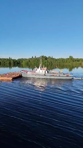 A boat in a body of water is seen from a high angle. The boat is gray and has a small crane on the back. It is pulling a small brown barge behind it. The water is dark blue and has small waves. The sky is clear and light blue. Green trees line the distant shore.