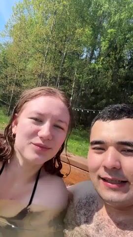 The video shows a young couple in a hot tub outdoors. The woman has light skin, wet reddish-brown hair, and wears a black bikini top. The man has dark hair, medium skin tone, and appears to be shirtless. They are close together, smile at the camera, kiss briefly, and then smile again. The background shows trees, a blue sky, and some overhead string lights. The hot tub water is murky.