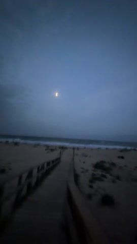 The video shows a beach at dusk or night. The sky is cloudy and dark. A person in a red hooded jacket walks on a wooden boardwalk that leads toward the ocean. The beach is sandy with some vegetation. The ocean waves are visible in the distance. The wind is loud.