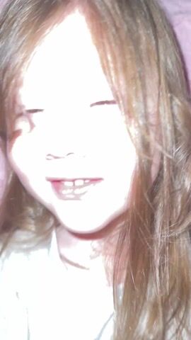 A close-up video shows a young girl with light brown hair and brown eyes, smiling at the camera. She wears a gray shirt and a thin gold necklace. Her hands are raised, partially obscuring her face at times. The background is blurred, showing a pink surface. The girl speaks, but the audio is unclear.