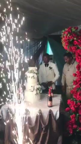 The video shows a wedding reception, with a groom in a white suit standing behind a four-tier white wedding cake, preparing to cut it. There is a bottle of champagne on the table next to the cake. A sparkler display is emitting sparks in front of the table. Red flower arrangements are visible on either side of the scene. Another person, likely a guest, is partially visible on the right, and other people are blurred in the background to the left. The event is taking place under a tent or canopy with draping fabric.