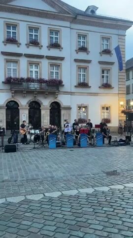 A street band performs in front of a building. The band consists of a keyboard player on the left, a guitarist and drummer in the center, and several horn players to the right. The building behind them has a light-colored facade, multiple windows with flower boxes, and a balcony. A cobblestone plaza is in the foreground. The scene appears to be taking place during the day, possibly late afternoon or early evening, given the lighting.