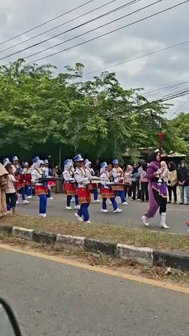 A marching band comprised of young people in uniform walks down a street. The band members wear white shirts, blue vests, and red skirts or pants, with blue and white hats. Some carry musical instruments, while others have what appear to be decorative staffs or batons. On the side of the road, a crowd of onlookers watches the procession. Some of the spectators are holding umbrellas to shield themselves from the elements. The environment appears to be outdoors, with trees and power lines visible in the background. The sky is overcast.