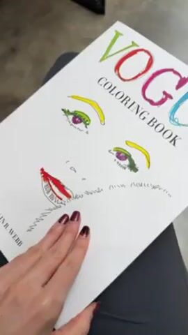 A hand holding a Vogue Coloring Book, with the cover facing up and slightly tilted. The book has a simple illustration of a face on the cover, with colorful eyes and lips. The hand opens the book, revealing pages filled with black and white fashion illustrations of women in stylish outfits. The hand continues to flip through the pages, showing more drawings of women in different poses and clothing. The background is blurred.