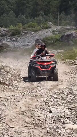 This video captures an ATV riding on a dirt path through a mountainous, forested area. The ATV is red and black, with a rugged design suitable for off-road terrain. Two people are riding on the ATV, with the person in the front steering and the person behind them holding up a smartphone, possibly recording or taking a photo. In the background, several other individuals are visible, also appearing to be on a dirt path or trail. The environment is characterized by rocky terrain, scattered boulders, and dense green foliage covering the hillsides. The lighting suggests it is daytime with clear, sunny conditions, casting shadows on the ground. The overall scene depicts an adventurous outdoor activity in a natural, somewhat remote landscape.