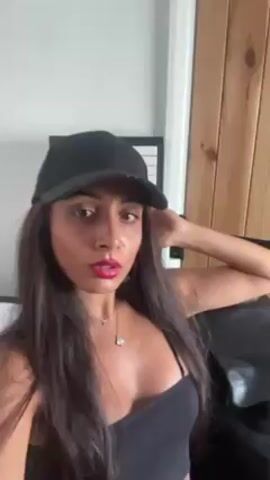 A woman with long brown hair wearing a black tank top and black baseball cap is sitting on a black couch. She is facing the camera and turns her head slightly to the side at the end of the video to show a piano keyboard art piece hanging on the wall. The wall is white with a light brown wooden door on the right. The woman is wearing bright red lipstick and a silver necklace.