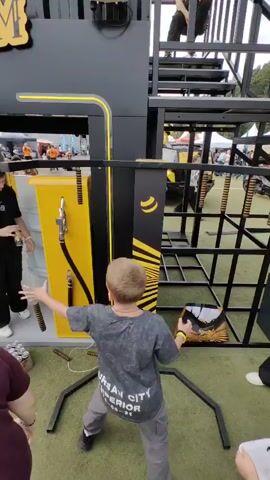 A young boy with blond hair stands in front of a yellow and black structure designed to resemble a gas pump, with a hose attached. He is wearing a grey t-shirt with text on it and grey pants. His arms are raised, and he appears to be interacting with a game or activity. To his left, a person wearing a black t-shirt is crouching near a stack of cans. To his right, a black metal cage-like structure is visible. The ground is covered in green artificial turf. In the background, tents and other structures are present, suggesting an outdoor event. The lighting indicates it is daytime.