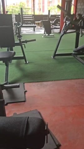 The video pans across an outdoor gym, revealing various weightlifting machines on a floor of green turf. Adjacent to the turf is a section of red flooring with black mats. The gym equipment includes a preacher curl machine, a leg extension machine, and a leg curl machine. There are also various weights and benches. A red punching bag hangs from the ceiling. Outside the gym, a view of urban buildings is visible.