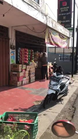 A person stands in front of a storefront, organizing merchandise. The storefront is lined with shelves holding various products, including bags of snacks and boxes of packaged goods. Several motorcycles are parked outside the storefront. The scene appears to be a commercial street with buildings and signage visible in the background. Sunlight suggests it is daytime.