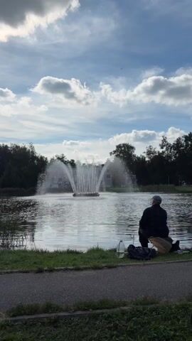 A person is fishing in a pond with a fountain in the background. The person is seated on the grassy bank of the pond, facing away from the camera, and holding a fishing rod. There are some bags and a bottle next to the person. The pond is surrounded by trees, and the sky is partly cloudy.