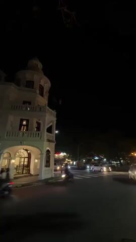 The video depicts a nighttime street scene.  A white, multi-story building with a domed top stands at the corner of the frame on the left.  The street is active with vehicles including motorcycles and cars, their headlights visible. Across the street, there is a park-like area with illuminated yellow structures, likely pavilions or gazebos. Trees line the street, and a black sky is visible above.