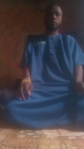 A man wearing a blue garment and a yellow bracelet sits cross-legged on the floor. The camera angle is looking down at him. The background is a wall. The man then bends forward, so the camera is focused on the back of his head.