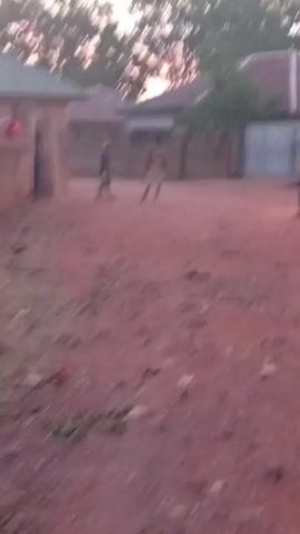 The video depicts a dusty, open area possibly in a village or rural setting. The ground is reddish-brown and appears to be made of dirt or clay. Several structures, possibly houses or buildings, surround the open area. There are people walking in the background, appearing to be children or young adults. The lighting suggests it might be early morning or late afternoon. There is also some sparse vegetation and debris scattered around. The video appears to be taken from a handheld device, and it's slightly shaky.