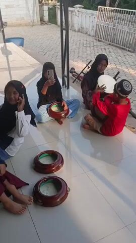 A group of young girls are sitting on a tiled floor. There are at least three girls visible, all wearing hijabs. Two of the girls are sitting cross-legged, while one is leaning back on her hands. In the foreground, there are two round, wooden tambourines with metal jingles. One girl on the right is holding a tambourine and a drumstick. Another girl on the left is holding a phone and appears to be taking a picture or recording a video. In the background, there is a large bass drum. The scene appears to be outdoors, possibly on a patio or porch, with a white building and fence visible. The lighting suggests it is daytime, possibly late afternoon.