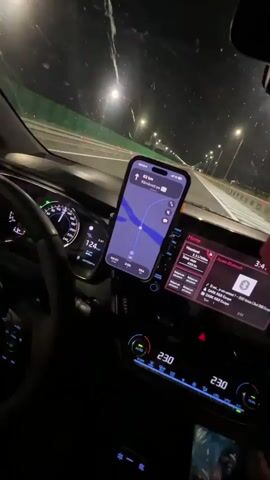 The video shows the interior of a car at night, driving on a highway. The driver's side of the car is visible, with the steering wheel and dashboard. A smartphone is mounted on the dashboard, displaying a navigation app with a map and route. The car's speedometer shows a speed of 125-127 km/h. The central infotainment screen is also visible, displaying the time as 3:46. The climate control display shows a temperature of 23 degrees Celsius. Outside the car, streetlights illuminate the highway, and guardrails are visible. The video appears to be a first-person perspective from the driver's seat, with a hand visible on the steering wheel at one point. The audio consists of music and possibly some indistinct vocalizations.