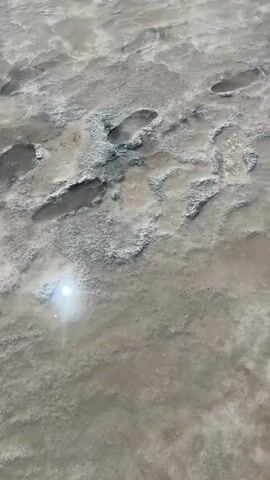 The video shows a close-up, overhead view of wet sand with footprints. There is a bright reflection of light in a puddle of water on the sand. The footprints are partially filled with water and appear to be from a person walking. The sand is a light brown color, and the water is clear.
