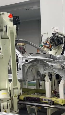 The video shows an automated car manufacturing process. Several industrial robotic arms are positioned around the chassis of a car. The chassis is mounted on a platform that appears to be moving along a production line. The robotic arms are equipped with various tools and appear to be engaged in assembly or welding operations on the car's frame. The environment is a brightly lit factory floor with large white walls and industrial equipment. There are no people visible in the video. The robots are the primary actors, moving precisely and efficiently to construct the vehicle. The focus is on the mechanical and automated nature of the car manufacturing process.