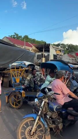 The video shows a street scene with several motorbikes and three-wheeled vehicles. In the foreground, a person wearing a pink shirt and a black baseball cap sits on a blue motorcycle. To the left of the motorcycle, a person in a light blue shirt sits on a blue three-wheeled vehicle. Behind them, another person in a camouflage jacket sits on a blue three-wheeled vehicle. In the background, there is a truck with a large black tarp, and several buildings line the street. There is also an ice cream cart with pictures of different flavors of ice cream. The sky is blue with some white clouds.