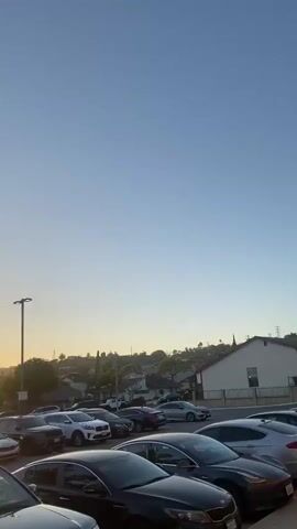 The video begins with a wide shot of a clear blue sky over a suburban neighborhood with houses and trees. A row of parked cars is visible in the foreground. The camera then pans to the left, revealing a sunset with the sun peeking through the trees, casting a warm glow on the sky. The camera continues to pan, showing more cars parked in what appears to be a parking lot. It then moves to a closer view of the cars, highlighting their various models and colors. The video shifts to show the exterior of a building with a brick facade and a large entrance with green and white doors. A sign above the entrance reads "NO SKATEBOARDING, ROLLERBLADING, OR SCOOTERS ALLOWED AT ANY TIME." The camera then focuses on the entrance, showing a glimpse of a basketball court inside where people are playing. Finally, the video shows a close-up of the brick wall and the tiled decorative band above the entrance.