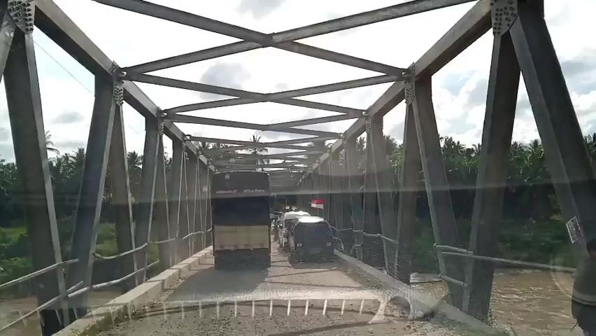 The video shows a first-person perspective from inside a vehicle driving across a metal truss bridge. The bridge is narrow, with a single lane of traffic. Several vehicles are on the bridge, including a large truck, a white van, and a black van. Some vehicles appear to be waiting. The road surface on the bridge is uneven and appears to be under construction or repair. The bridge spans a muddy river, and dense vegetation, including palm trees, lines the banks on both sides. The sky is overcast with scattered clouds.
