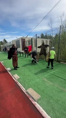 The video shows an outdoor reenactment scene, likely a Viking theme, where two individuals are engaged in mock combat with swords and shields on a green, artificial turf surface. Both participants are wearing protective headgear and casual sportswear. The combat area is bordered by a red running track, a chain-link fence, and a building in the background with light-colored paneling. A bear skin rug hangs from the fence, adding to the theming. Two other individuals, dressed in what appear to be Viking-era costumes, stand nearby, seemingly supervising or observing the mock battle. There is a table with items on it between the individuals in costume. The weather appears sunny with a blue sky dotted with clouds.